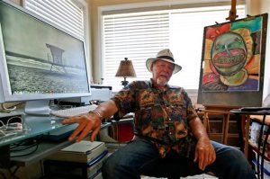FILE - This July 16, 2014 file photo, artist Michael Gross sits in his studio between one of his paintings and one of his photos on his computer screen in Oceanside, Calif. Gross, an artist who created two of the most distinctive pop culture images of the 20th century, died Monday, Nov. 16 2015. He was 70. Gross first gained wide attention in 1973 for the National Lampoon cover of a dog with a gun to its head and the words, "If You Don't Buy This Magazine, We'll Kill This Dog." A decade later, he created the enduring symbol of a confused looking ghost in the middle of a slashed red circle for the film "Ghostbusters." (AP Photo/Lenny Ignelzi, File)